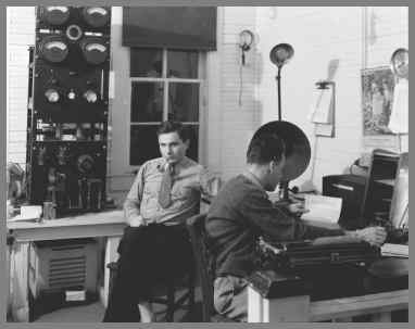 WUG2 radio room with transmitter in background and one operator relaxing with a pipe and another at a desk on the right pounding the key.