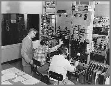 Two operators sitting in front of equipment rack panels at small rack panel mounted desks. Supervisor standing on the left.