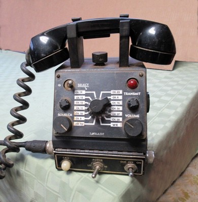 A sloped front control head with handset on hanger on the top, a 14 channel selector swithg on the slope along with volume and squelch knobs and 6 other lights and toggle switches.