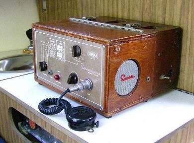 Tabletop Radio transceiver with a hinged top, 3 small knobs and a speaker grill on right side. Mmicrophone laying in front.